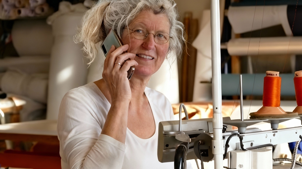 Een vrouw met bril en grijs haar die lachend aan het bellen is bij een naaimachine in een atelier.