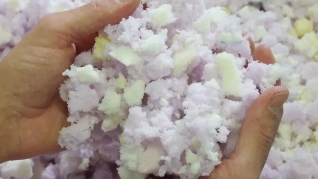 Close-up van twee handen die een grote hoeveelheid wit en lichtpaars gemalen schuimrubber vlokken vasthouden voor kussenvulling.
